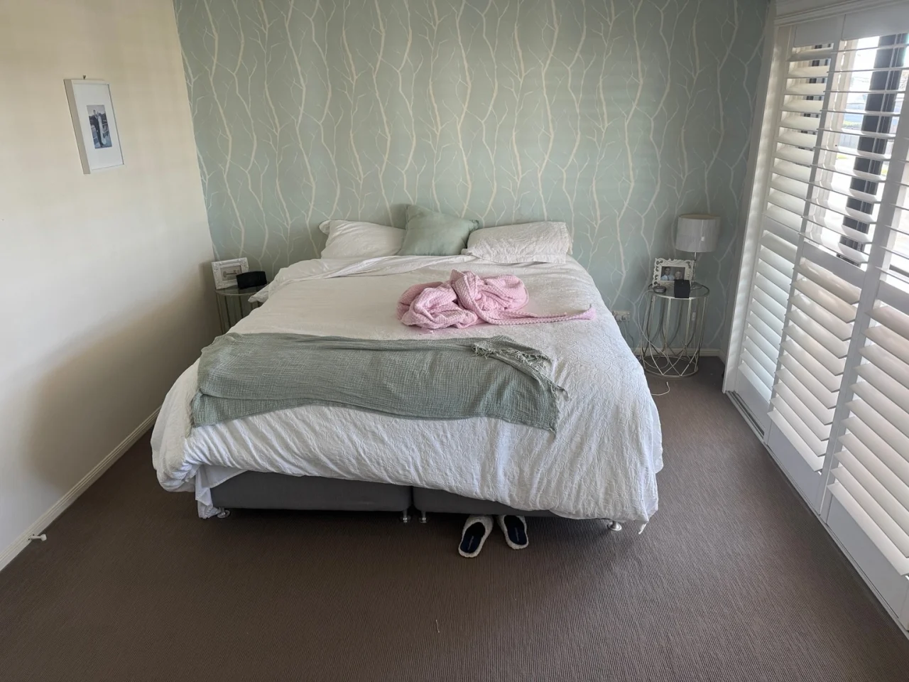 A real bedroom as photographed on a phone: an unmade bed, two side tables, slippers on the carpet.